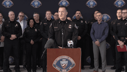 A uniformed fire official from San Bernardino County Fire Department stands at a podium during a press conference, speaking into microphones labeled with Telemundo and other news outlets. He is surrounded by a group of men and women, many of whom are also in fire department uniforms, standing against a backdrop featuring the San Bernardino County Fire Department emblem. The podium displays the department's badge with the words "San Bernardino County Fire - Rescue - EMS." The setting suggests an official announcement or briefing.